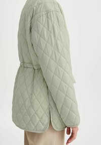 Chaqueta acolchada de color verde claro con cintura ceñida, que presenta un dobladillo redondeado y un patrón de diamante diagonal en la tela.