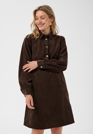 Femme souriante, portant une robe en velours côtelé marron à manches longues avec des boutons, des créoles dorées et un bracelet, debout devant un fond uni.