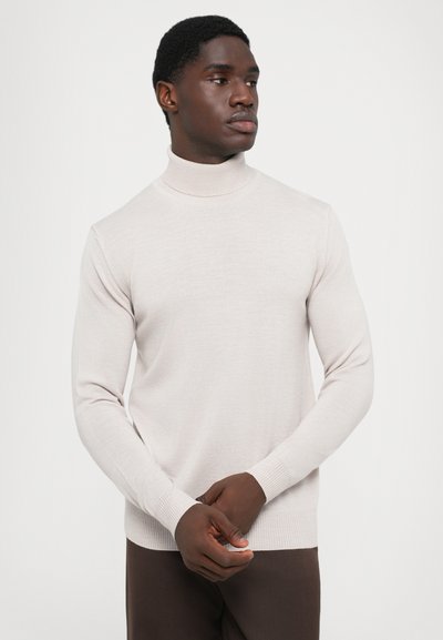 Maglione girocollo beige chiaro realizzato in tessuto morbido e texturizzato, silhouette aderente, con polsini e orlo a coste. Indossato con pantaloni scuri.