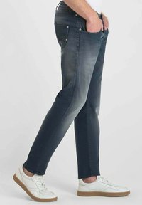 Jeans de color azul oscuro con un acabado desgastado, diseño ajustado, estilo de cinco bolsillos, material de algodón, y combinados con zapatillas blancas.