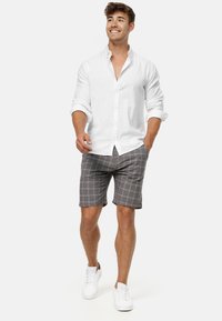 Chemise en lin blanche à manches retroussées associée à un short à carreaux gris. Modèle portant des baskets blanches, présentant une tenue d'été décontractée.