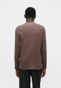 Mand set bagfra iført en tekstureret brune langærmet skjorte og løse sorte bukser mod en ensartet lys baggrund.