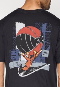 Μαύρο βαμβακερό t-shirt με γραφικό σχεδιασμό ενός χεριού που κρατά μια κόκκινη μπάλα μπάσκετ στην πλάτη, με αφηρημένα μπλε και γκρι στοιχεία και το λογότυπο της Nike.