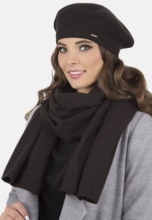 Czarna dziana beretka i dopasowany szalik wykonany z miękkiego materiału. Szalik elegancko opada na szary płaszcz. Minimalistyczny akcent na beretce.