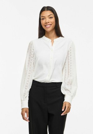 Frau lächelnd, trägt eine weiße Bluse mit Knöpfen und langen Spitzenärmeln sowie schwarze Hosen, steht vor einem schlichten weißen Hintergrund.