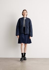 Marinblå quiltad jacka med rund hals, bärs över en randig topp och en mörk kjol. Svarta platåskor och ankelsockor fulländar outfiten.