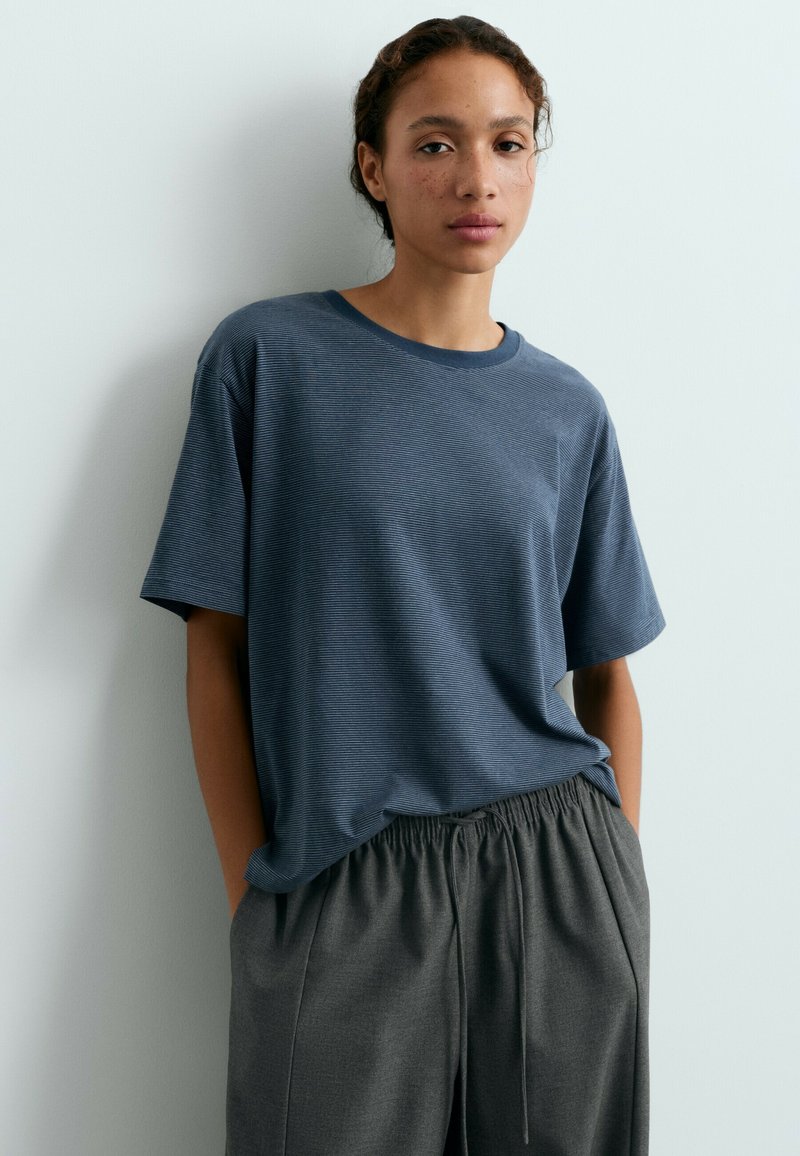 Jeune femme aux cheveux bouclés portant une chemise bleue ample à manches courtes et un pantalon gris à taille élastique, debout contre un mur blanc.