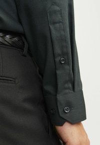 Camisa de vestir de patrón verde oscuro con puño estrecho, que cuenta con dos botones negros y un cuello estructurado. Combinada con pantalones negros.