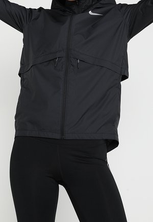 Person trägt eine schwarze Nike Jacke mit Reißverschluss und Kapuze sowie schwarze Leggings und hebt die Arme vor einem einfarbigen hellen Hintergrund.