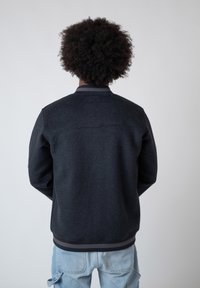 Person with curly hair wearing a dark gray bomber jacket and light blue jeans, standing facing a plain light gray background.