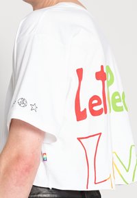 Vit bomullströja med färgglada textgrafiker i rött, grönt och gult. Korta ärmar med en regnbågsaccent vid fållen.