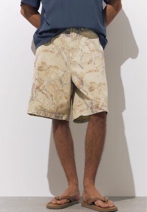 Muškarac u bež kratkim hlačama s cvjetnim uzorkom, tamnoplavoj košulji i smeđim japankama stoji uz bijeli zid.