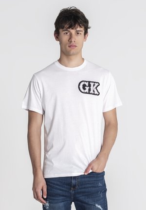 Camiseta de algodón blanca de manga corta, que presenta un logo negro "GK" en el lado izquierdo del pecho y un cuello redondo. Combinada con jeans azules desgastados.