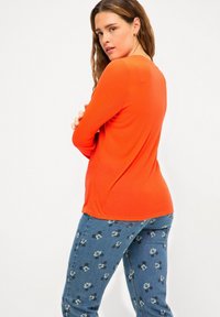 Top a maniche lunghe a coste di un vivace arancione, abbinato a jeans in denim blu con un motivo di Topolino, visto da un leggero angolo.