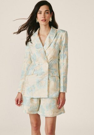 Mujer con cabello largo y oscuro que lleva una chaqueta de traje crema y pantalones cortos a juego con un estampado floral azul y pendientes blancos.