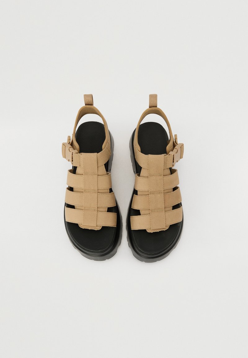 Beige Riemchensandalen mit schwarzen gepolsterten Sohlen und verstellbaren Knöchelriemen, von oben auf einem weißen Hintergrund gezeigt.