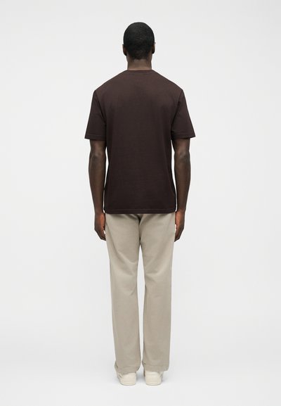 Άνδρας που στέκεται γυρισμένος από την πλάτη, φορώντας ένα σκούρο καφέ κοντομάνικο πουκάμισο, μπεζ παντελόνια και λευκά αθλητικά παπούτσια, μπροστά σε απλό ανοιχτό φόντο.