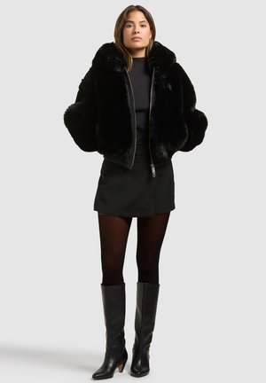 Femme debout portant une veste en fausse fourrure noire, une jupe courte noire, des collants noirs et des bottes en cuir noires montant jusqu'aux genoux sur un fond uni.