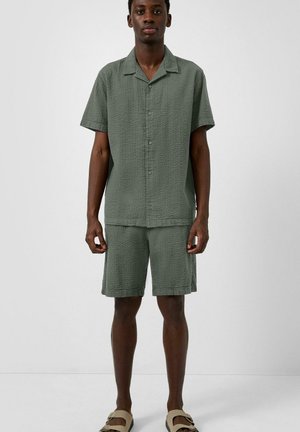 Man draagt een gestructureerd olijfgroen overlopen kort mouwens overhemd en bijpassende shorts met beige sandalen, staand tegen een effen achtergrond.