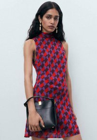 Kurzes Kleid mit einem geometrischen roten und lila Muster, hohem Kragen, ärmellosem Design und einer schwarzen Handtasche, die in der linken Hand gehalten wird.