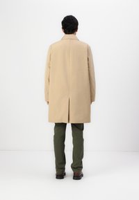 Palton bej cu vent posterior, mâneci lungi și croială dreaptă, asortat cu pantaloni verzi și pantofi negri. Design minimalist, fără modele vizibile.