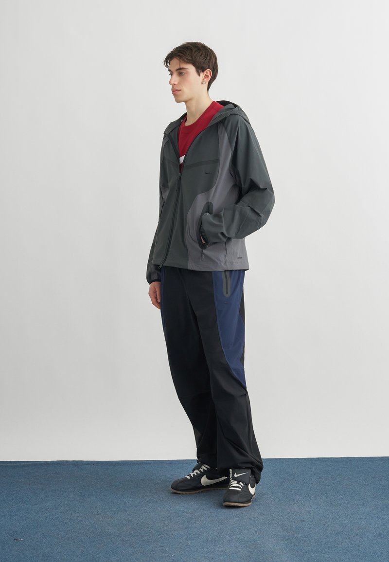 Junger Mann steht und trägt eine graue Kapuzenjacke, ein rotes Hemd, schwarze und blaue Hosen sowie schwarze Nike-Sneaker auf einem blauen Teppich.