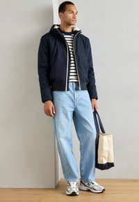 En marineblå zip-jakke med en blød inderside, en stribet t-shirt under, lyseblå jeans og grå sneakers. En canvas tote-bag i hånden.