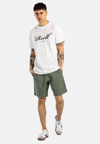 Camiseta de algodón blanca con "Reell" impreso en negro, pantalones cortos verdes oliva y zapatillas blancas con detalles en gris. El modelo está de frente.