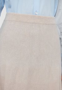 Vaaleabeige neulehame, jossa on pehmeä pinta ja leveä vyötärönauha, osittain piilossa vaaleansinisen kauluspaidan alla.