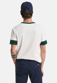 Men's short-sleeve, cream cotton t-shirt with green and navy striped cuffs and collar. Back view shows a simple, relaxed fit design.