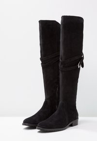 Botas negras de ante hasta la rodilla con un tacón plano, que cuentan con una cremallera lateral, correas decorativas y una textura suave.