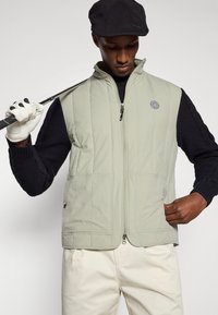 Ανοιχτό πράσινο γούνα είναι με ραφή και φερμουάρ μπροστά, διαθέτει δύο μεγάλα τσέπες και λογότυπο. Φορεμένο πάνω από μαύρο πουκάμισο με πλέξη καλωδίου μακρυμάνικο, συνδυασμένο με κρέμα παντελόνι.