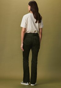 Mörkgröna kordflandyxor med bakfickor, kombinerade med en ljusgrå t-shirt. Modellen ses bakifrån mot en dämpad bakgrund.