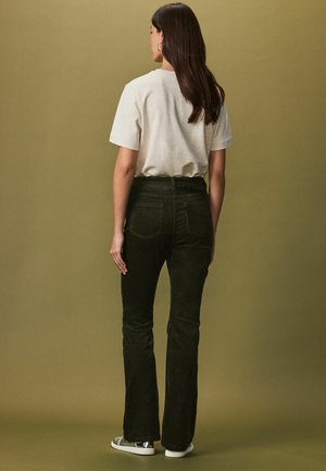 Mörkgröna kordflandyxor med bakfickor, kombinerade med en ljusgrå t-shirt. Modellen ses bakifrån mot en dämpad bakgrund.