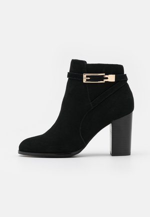 Bottine en daim noir avec talon bloc et une bride rectangulaire dorée autour de la cheville sur fond blanc.