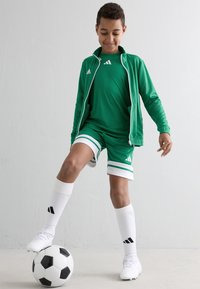 Pojke i grön fotbollsdräkt kontrollerar en svart och vit boll med foten mot en vit vägg.