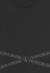 Czarna koszulka z okrągłym dekoltem, z poprzecznym paskiem z logo "CALVIN KLEIN JEANS" w odcieniach szarości na dolnej części przodu. Miękka w dotyku.