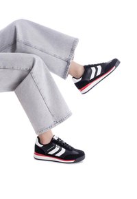 Zapatillas deportivas negras con rayas blancas, acentos rojos y suela de goma. Llevadas con pantalones de pierna ancha en gris claro. Diseño plano con cordones.