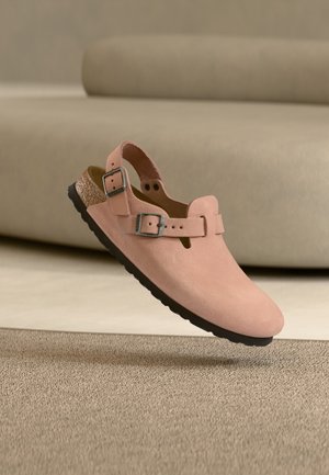 Scarpa slip-on in camoscio rosa tenue con due fibbie metalliche regolabili e sottopiede in sughero, mostrata su uno sfondo beige.