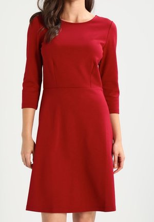 Frau trägt ein knielanges, eng anliegendes rotes Kleid mit dreiviertel Ärmeln und rundem Ausschnitt, steht vor einem schlichten hellen Hintergrund.