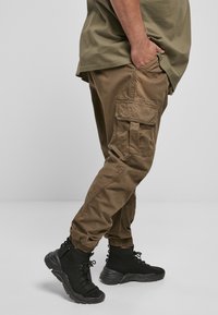 Cargo-byxor i brunt tyg med sidfickor, elastiska muddar och en bekväm passform, kombinerade med svarta höga sneakers.