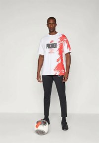 Balta Nike marškinėliai su raudonu grafiniu dizainu, juodu tekstu ir logotipu ant priekio; derinami su juodomis kelnėmis ir juodais batais, su futbolo kamuoliu.