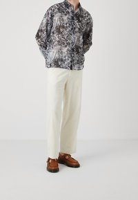Camicia a maniche lunghe nera e grigia con pattern, colletto alla cinese, abbinata a pantaloni a gamba larga color crema e scarpe di pelle marroni.