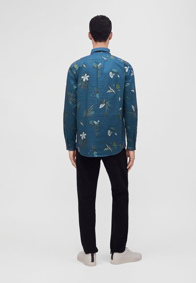 Homme portant une chemise à manches longues fleurie couleur sarcelle, pantalon noir et baskets blanches, debout dos à nous sur un fond clair uni.