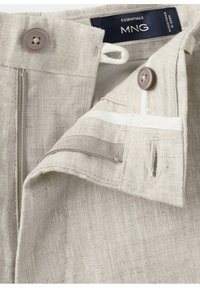 Zbliżenie na pas beżowych lnianych spodni, pokazujące dwa beżowe guziki, częściowy suwak oraz granatową metkę z napisem Essentials MNG Made in Bangladesh.