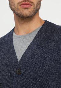 Marinblå stickad cardigan med V-ringning, prydd med en enda mörk knapp. Under finns en ljusgrå T-shirt synlig.