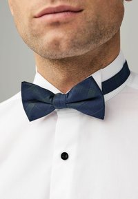Marineblau karierter Fliegen aus strukturiertem Stoff, getragen zu einem weißen Hemd mit einem schwarzen Knopf und schmalem Kragen.