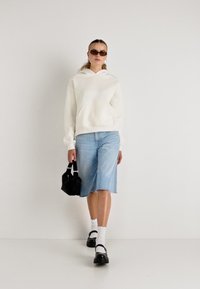 Felpa bianca con cappuccio, gonna in denim azzurro chiaro, borsa nera, scarpe nere e calzini bianchi. Gli accessori includono occhiali da sole scuri. Design semplice e casual.