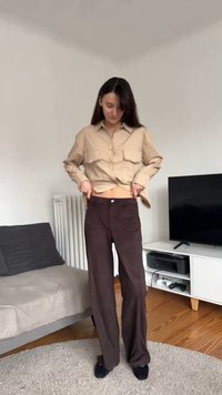 Camisa bege de manga comprida com dois bolsos no peito, combinada com calças largas castanhas de cintura alta. Tecido liso com um ligeiro brilho.