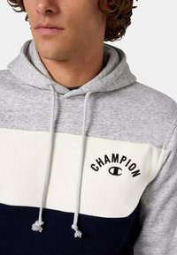 Mikina s kapucňou v šedej, bielej a námornej modrej farbe. Má jemnú textúru, vertikálne farebné bloky a vyšívané logo "Champion" na hrudi.
